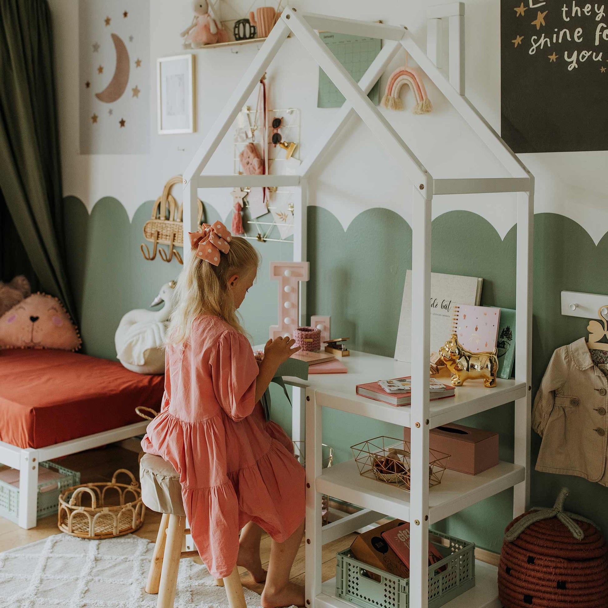 A young girl in a pink dress sits at a House-shaped toddler desk with built-in storage, deeply engaged in an activity. The beautifully decorated bedroom, filled with various toys, books, and a bed adorned with a stuffed animal, creates an ideal learning environment for a child.