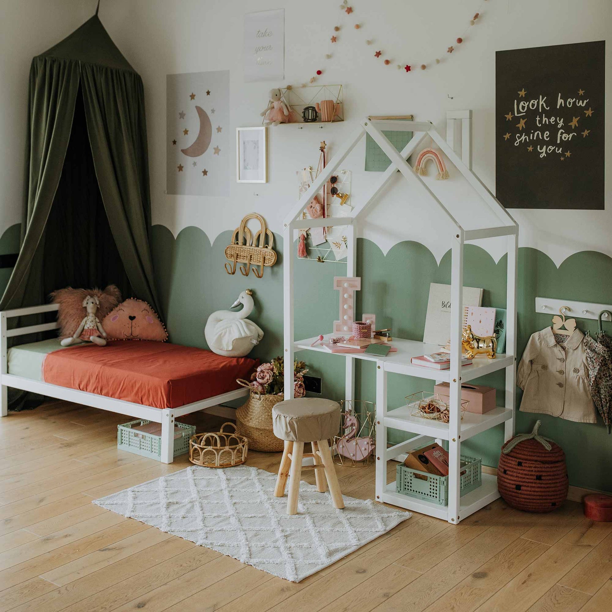 The children's room features a cozy arrangement with a bed, playhouse-style shelves, stools, and various toys. The soft green and white theme is accented by decorative wall art and a hanging garland. A House-shaped toddler desk enhances the child's learning environment. Light wooden flooring with a white rug completes the look.