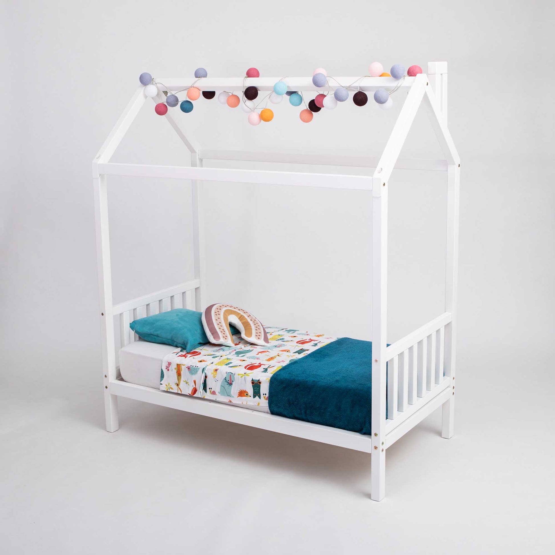 A white toddler house bed on legs with a headboard and footboard, with pom poms hanging from the ceiling.