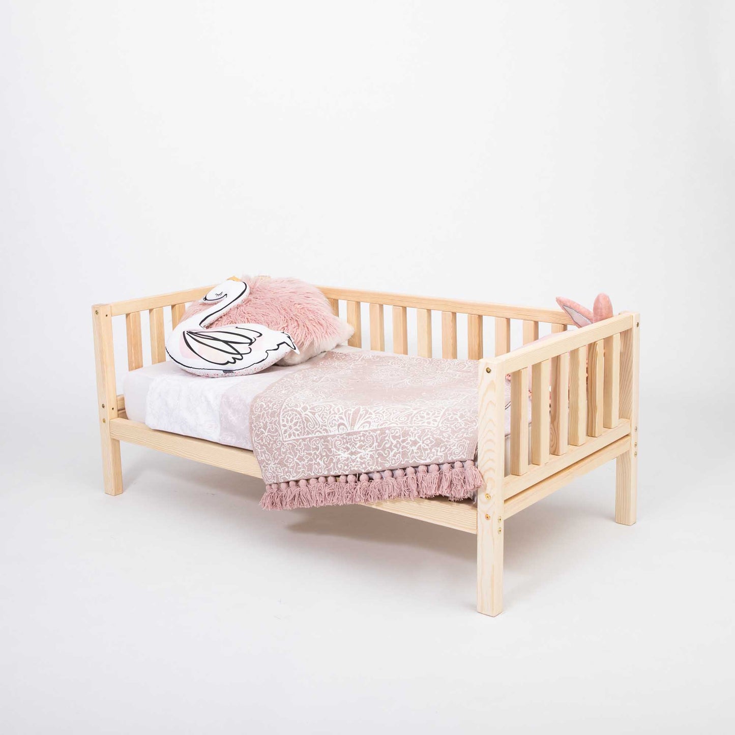 A Sweet Home From Wood 2-in-1 toddler bed on legs with a 3-sided vertical rail made of solid pine or birch wood, adorned with a pink blanket.