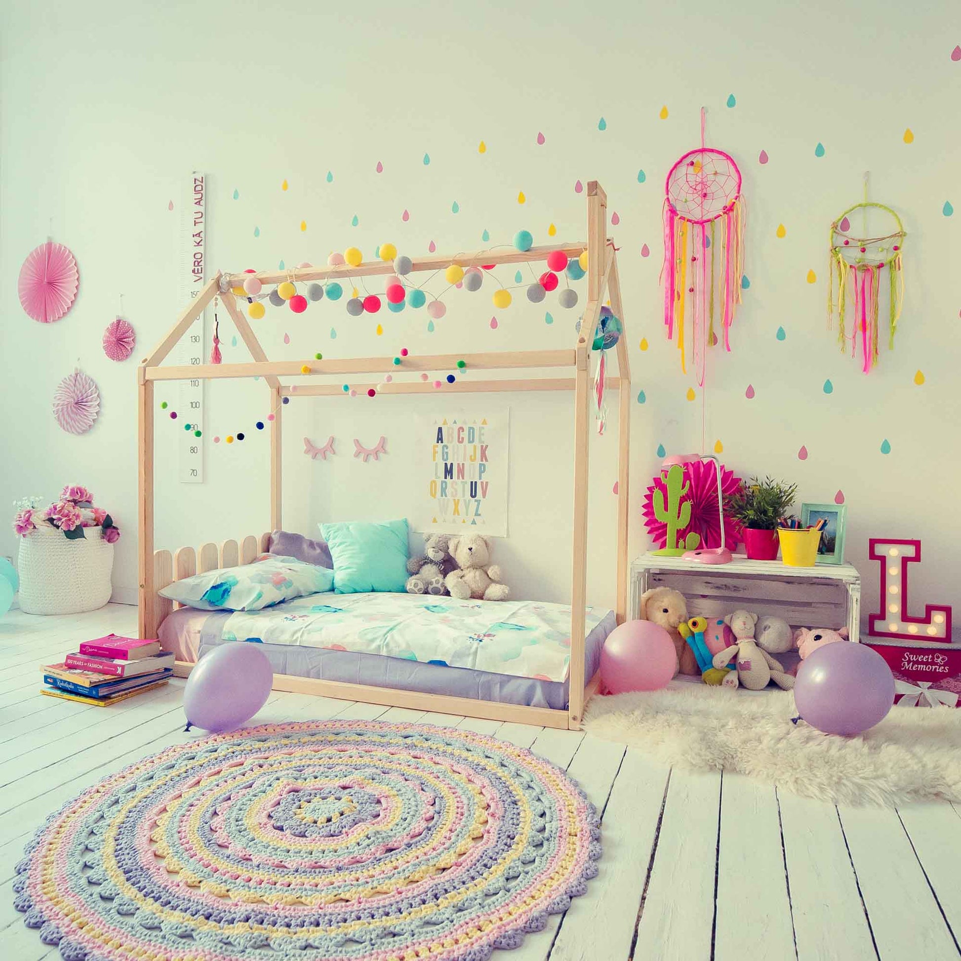 A children's bedroom featuring a Kids' house-frame bed with a picket fence headboard, stuffed animals, colorful wall decorations, a circular crochet rug, and books. Balloon decorations and dreamcatchers add a playful touch.