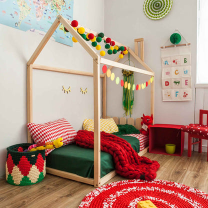 A children's room featuring a cozy Kids' house-frame bed with a picket fence headboard, adorned with green bedding, colorful pillows, charming wall decor, a red table and chair set, and a woven basket. A red and white circular rug adds a playful touch to the floor.