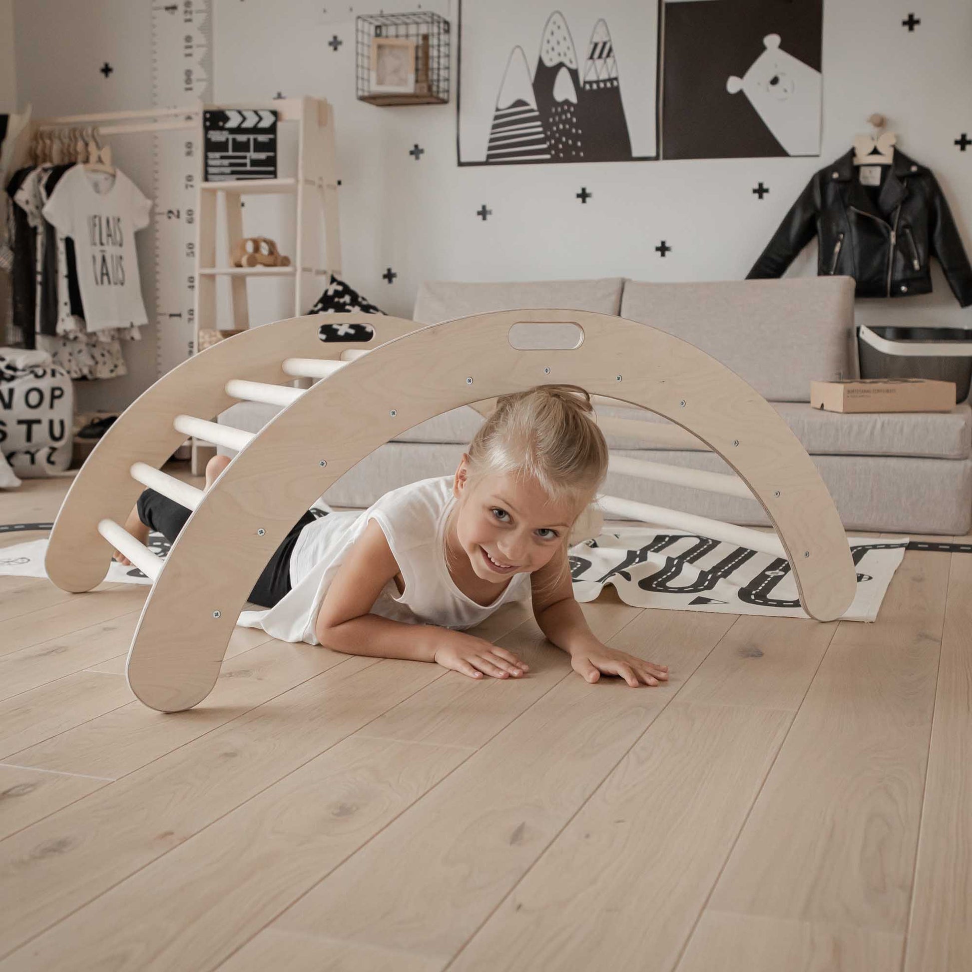 A little girl laying on the floor next to a wooden play structure and a Sweet Home From Wood climbing arch + Transformable climbing triangle + a ramp.