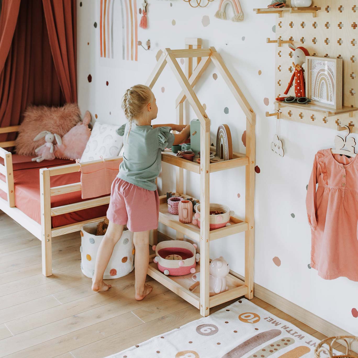 Types of toy shelves. A girl stanfing next to house shaped toddler toy shelf and playing.