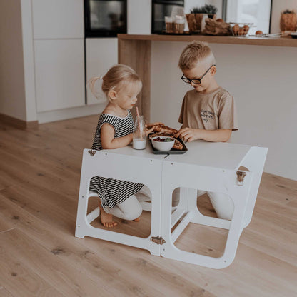 Two children using a 2-in-1 transformable kitchen tower - table and chair from Sweet Home From Wood to eat at a table in a kitchen.