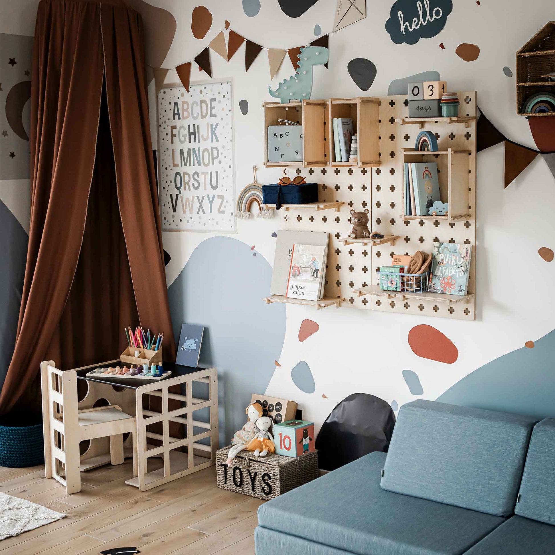 Child's room with educational and playful decor, including an alphabet chart, wooden shelves with toys and books, a small desk with art supplies, a toddler toy basket, and a blue sofa. Walls have abstract patterns. A 2-in-1 table and chair set or activity cube enhances the learning space.
