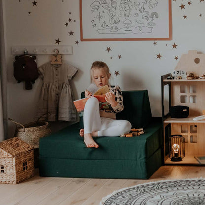 A little girl sitting on a green chair in a room with stars on the wall, surrounded by Sweet HOME from wood activity play couch set.