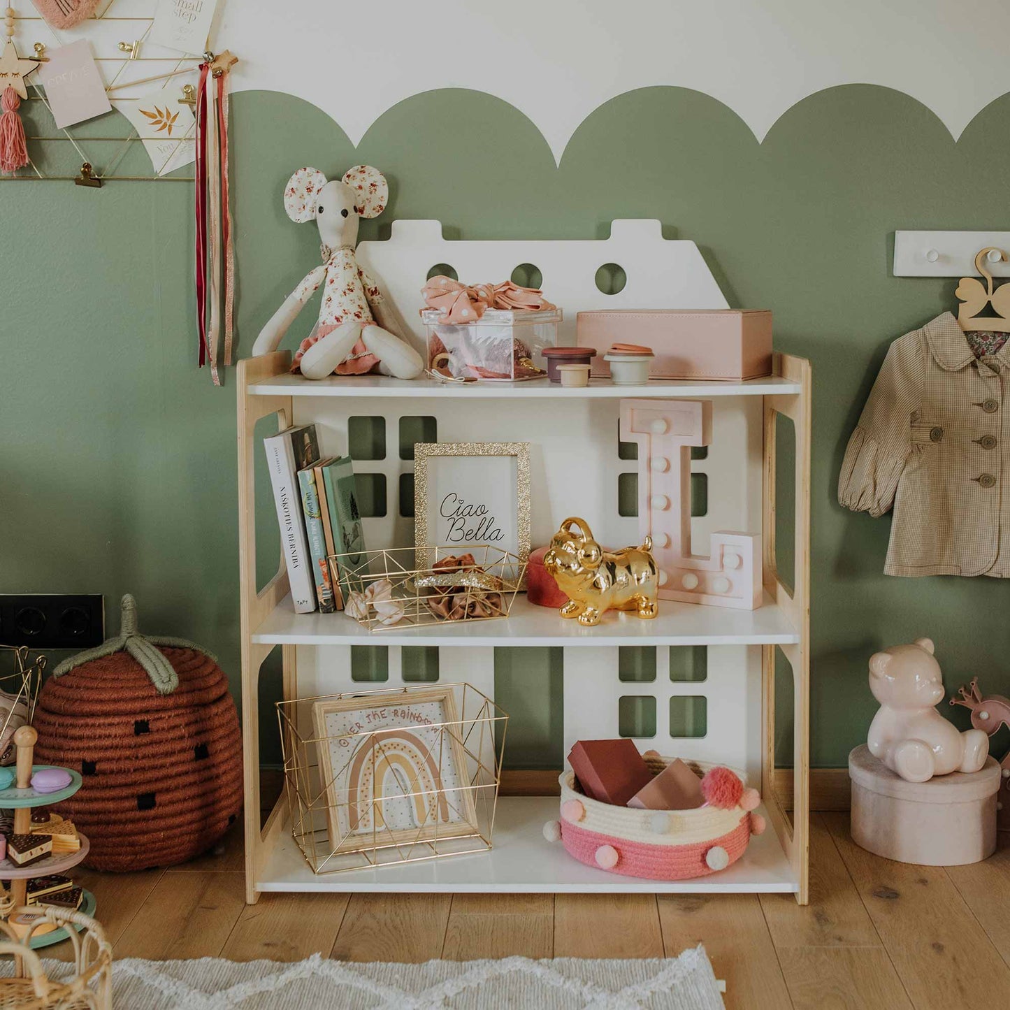 The 2-in-1 doll house and Montessori shelf in the children's room serves as both a toy display and an organizational solution, showcasing an assortment of toys and decorative items such as a stuffed mouse, golden bear figurine, books, and wooden blocks. The background wall is partially painted green, adding a cheerful touch to this kid's bedroom.