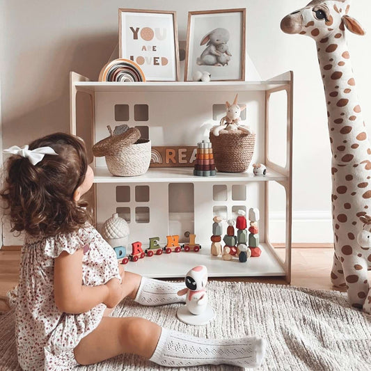 A little girl sits on the floor next to a 2-in-1 doll house and Montessori shelf.