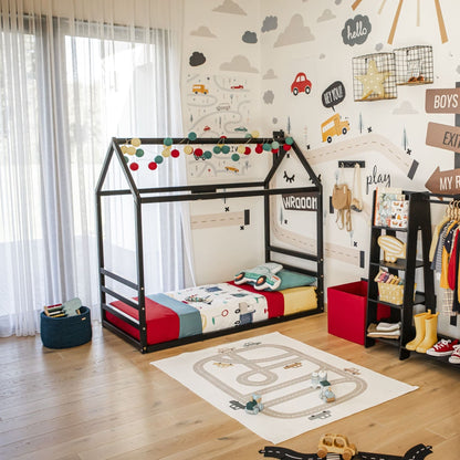 A children's room featuring a floor house-frame bed with a horizontal headboard and footboard, colorful bedding, a play mat adorned with a road design, and wall decorations showcasing cars and roads. Clothing and storage items are neatly organized around the room.