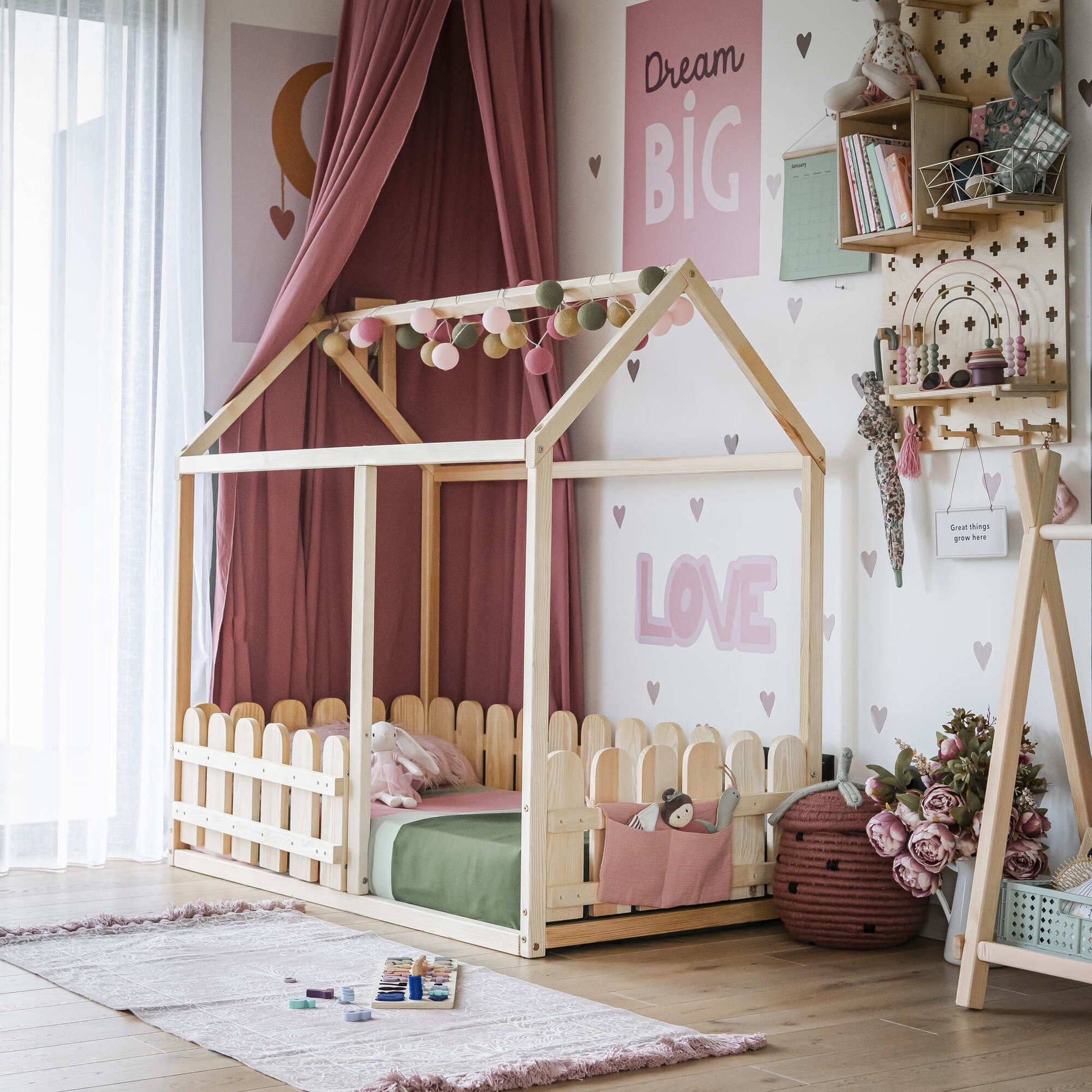 A children's room featuring a Platform house bed with a picket fence, complemented by a canopy and decorative lights. The walls are adorned with motivational posters, while a pink area rug lies beneath and a basket brimming with plush toys adds the finishing touch.