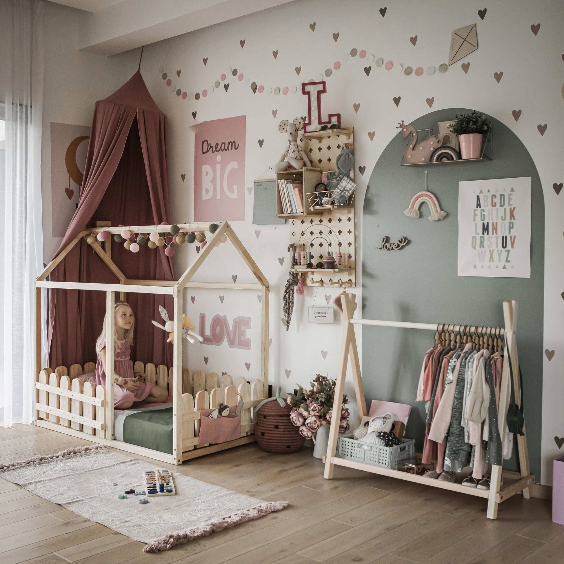 A child sits on a Platform house bed with a picket fence in a decorated room. The walls feature heart patterns, shelving, and various decorative items. A garment rack holding clothes is on the right side of the room.