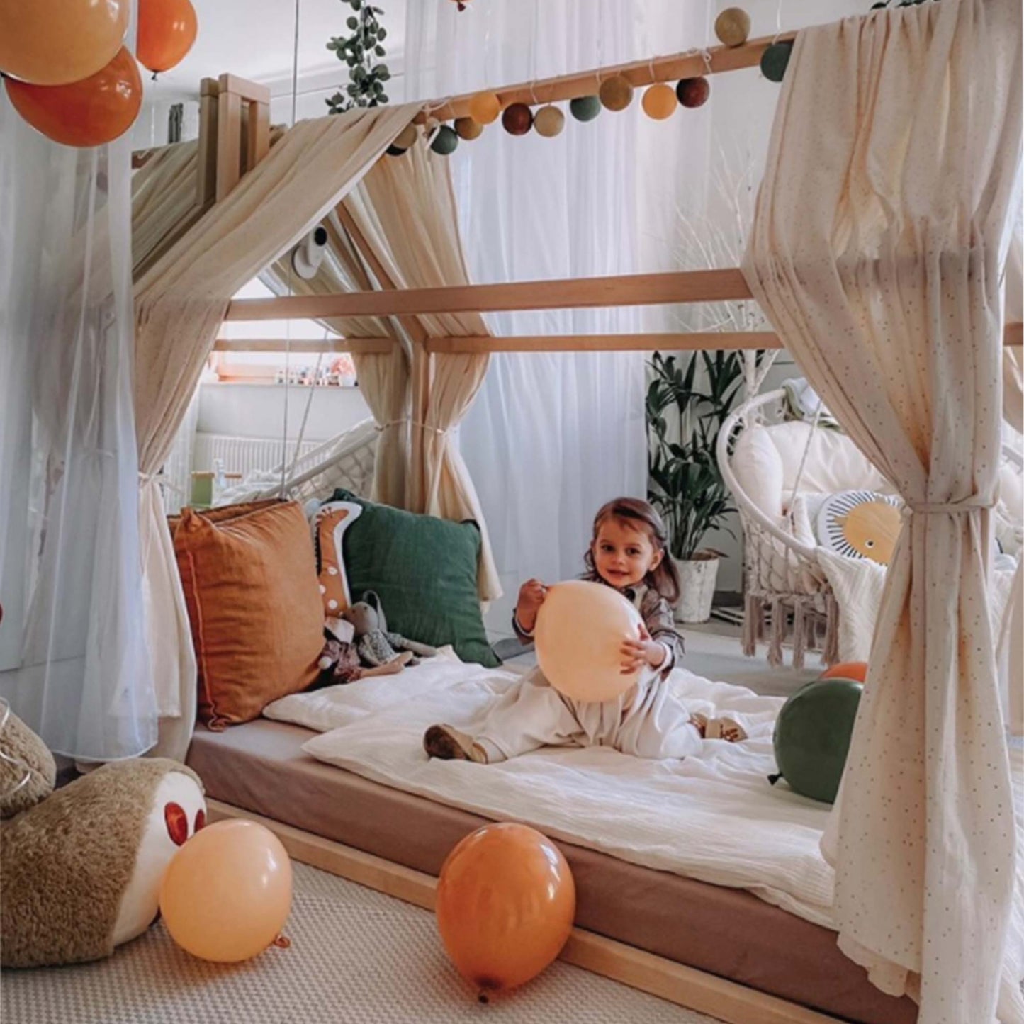 A little girl sitting on a cozy Sweet Home From Wood Wooden zero-clearance house bed with balloons and a house-shaped canopy.