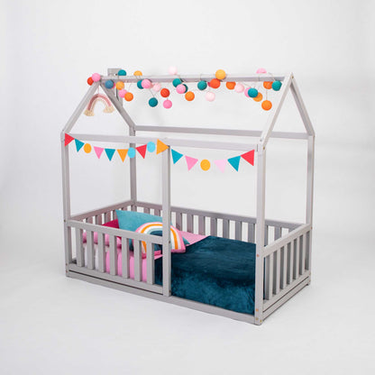 A cozy sleep haven for children in a Sweet Home From Wood Montessori floor house bed with rails and a colorful canopy with pom poms.