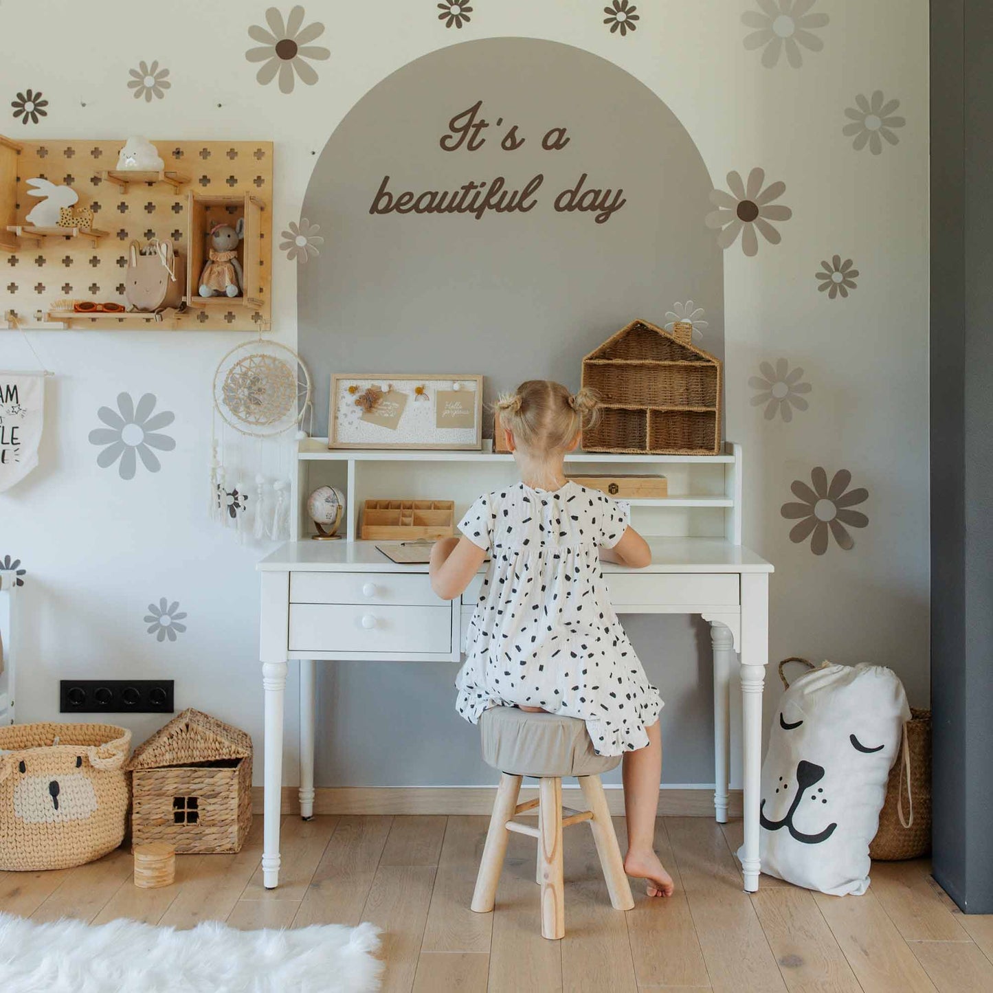 A child sits at a versatile pedestal desk adorned with decorative storage baskets and toys. The table with a hutch above provides extra space for books and art supplies. The wall behind features a decal reading "It's a beautiful day" adorned with floral designs.