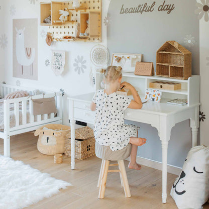 A child with blonde hair sits on a wooden stool, drawing at the versatile Table with a hutch, Pedestal desk in a children's room decorated with animal-themed items, wicker baskets, and a "beautiful day" sign on the wall. The desk features cable management and ample storage for all their art supplies.