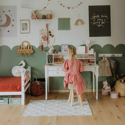 A child sits at a versatile, white table with a hutch and adjustable leg height in a neatly organized, pastel-toned room with green wall accents. The room features a small bed, various decorations, and storage baskets.