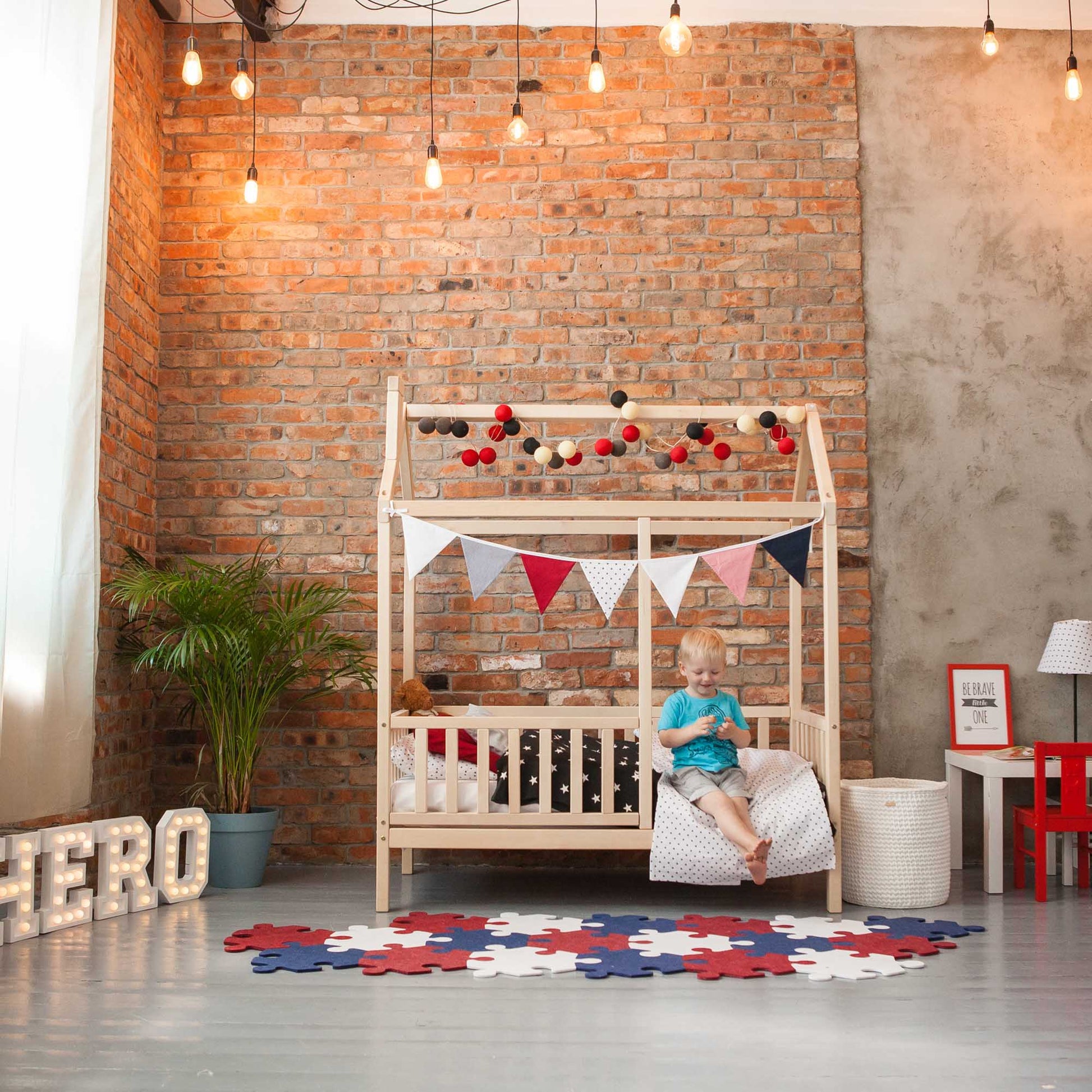 A child is sitting on a Kids' house bed on legs with a fence in a room with a brick wall.