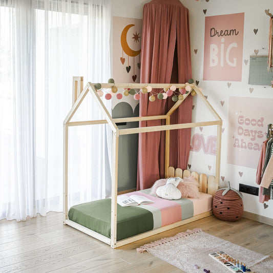 A children's bedroom with a Kids' house-frame bed featuring a charming picket fence headboard, adorned with string lights, colorful pillows, and a green and pink striped duvet. The walls are decorated with motivational posters. A basket and small shelf are situated nearby.