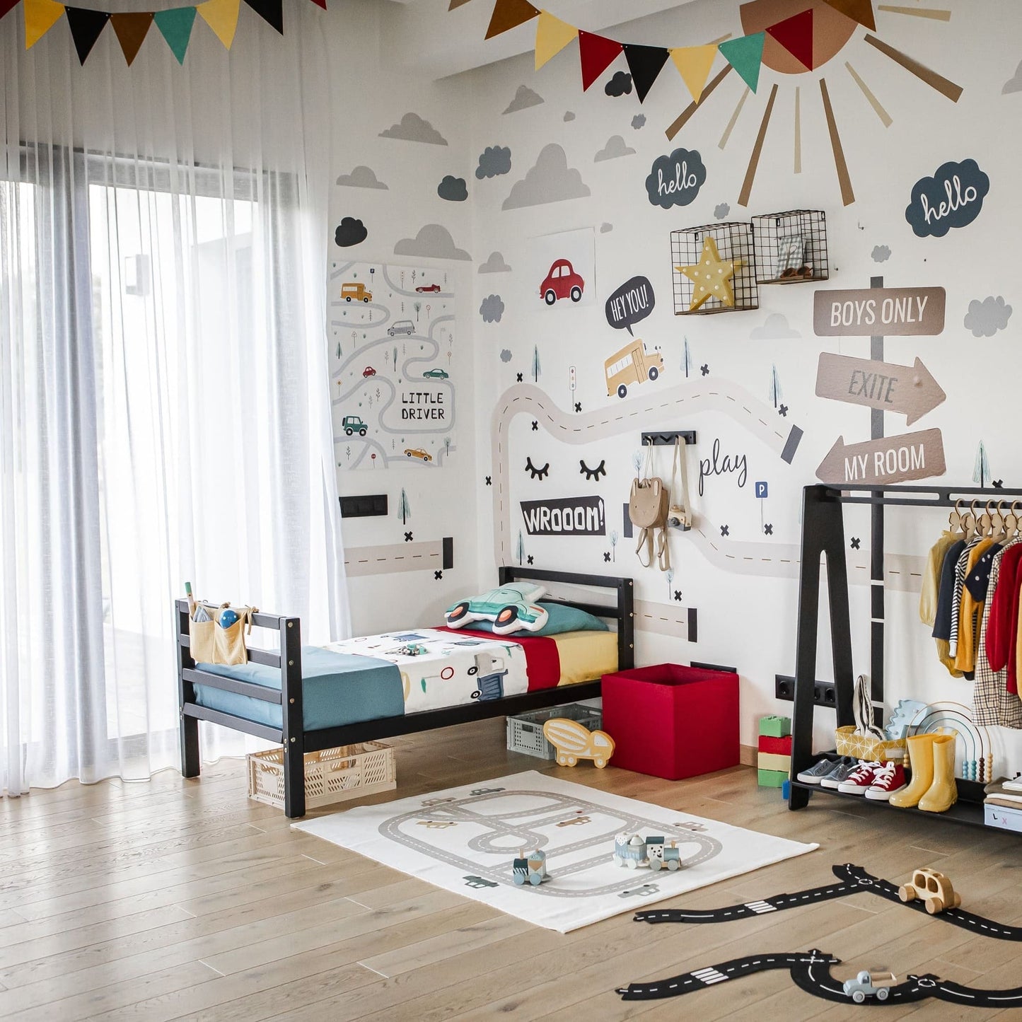 Children's bedroom with a vehicle-themed decoration. Includes a sturdy Montessori-inspired Kids' bed on legs with a horizontal rail headboard and footboard, play mat, race tracks, clothes rack, and colorful wall decals. Bright flags and sheer curtains add accents.