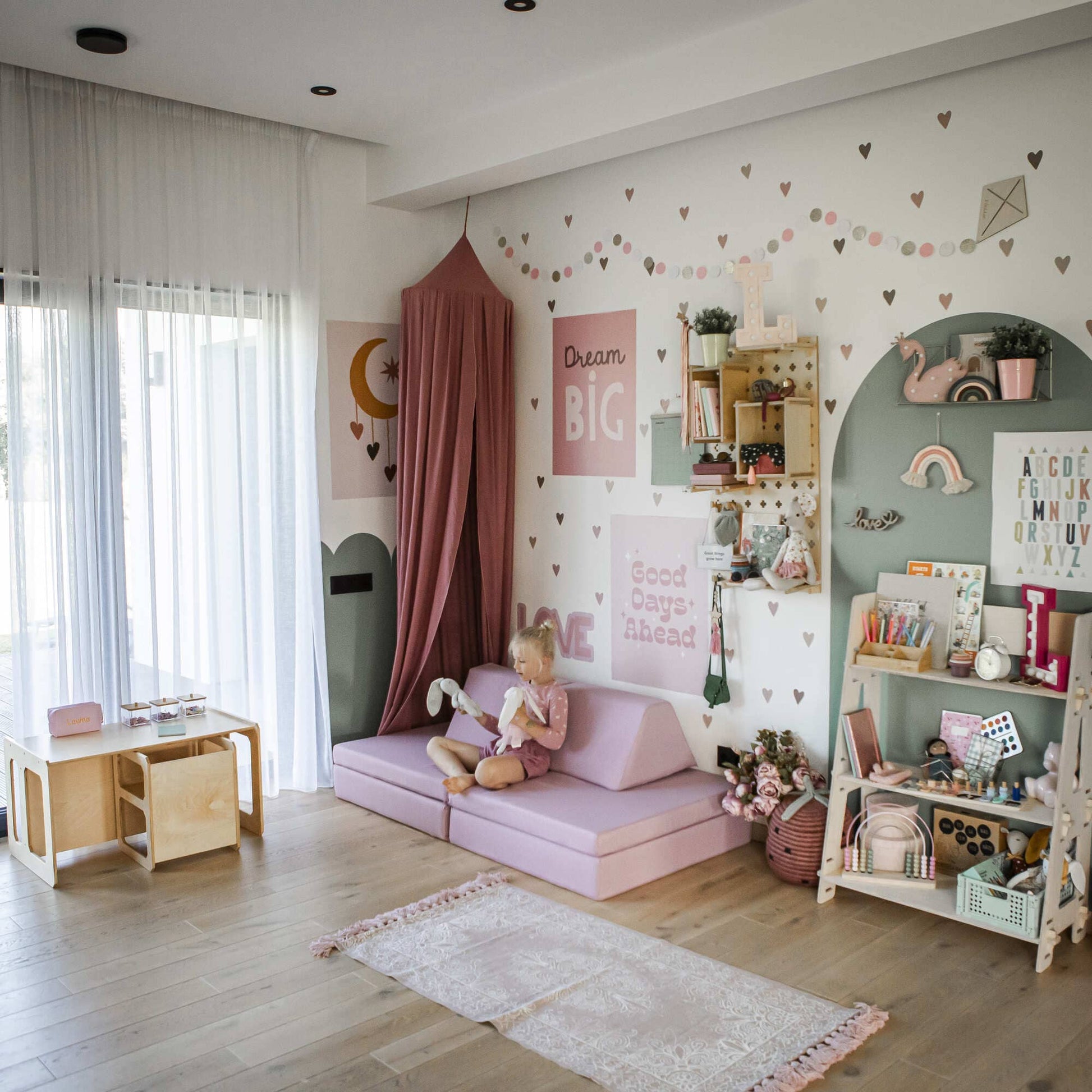 A child reads a book in a pastel-colored children's room with pink and green decor, enhanced by an Activity Play Couch Set that offers comfortable seating and play space, complemented by a small bookshelf, an indoor play gym area, and wall art featuring inspirational quotes.