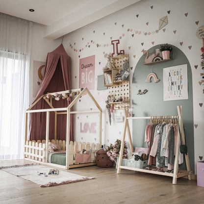 A children's room with a bed shaped like a house, a Kids' clothing rack with storage for easy kids storage solutions, books, and decorations on the wall. The decor features pastel colors and heart patterns. A window with sheer curtains is on the left, completing this cozy Montessori bedroom.