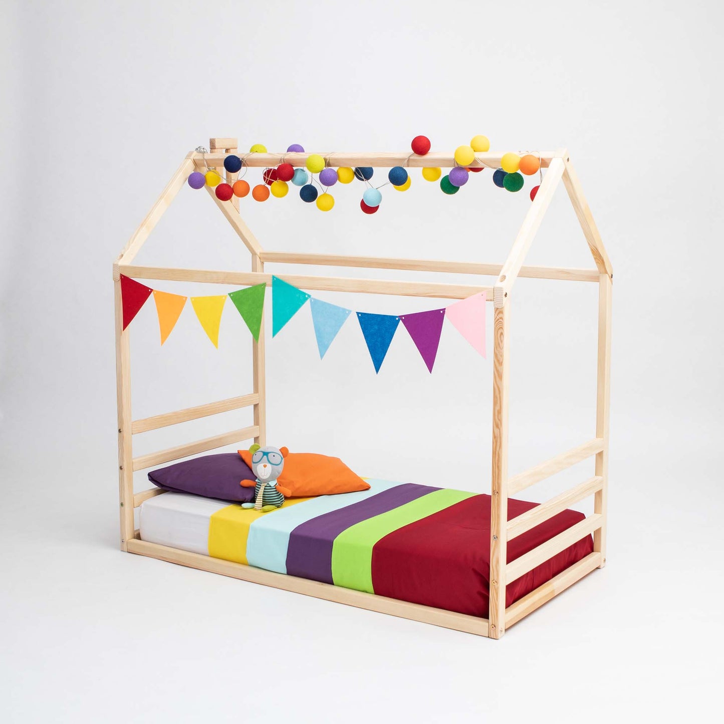 A floor house-frame bed with a horizontal headboard and footboard adorned with multicolored felt ball garland and triangular flags. The bed has colorful bedding consisting of striped blankets and pillows, and a plush toy sits on top.