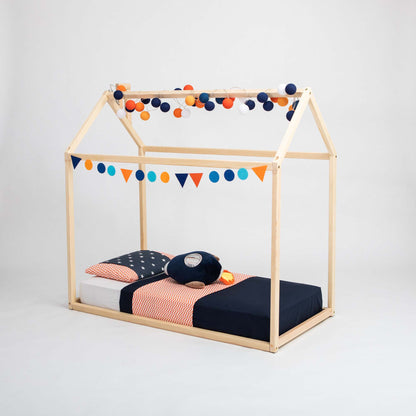 A Wooden zero-clearance house bed features a wooden house-frame design adorned with colorful bunting and string lights. This charming preschool bed is dressed in navy and orange themed bedding, perfectly blending style and functionality for the little ones.