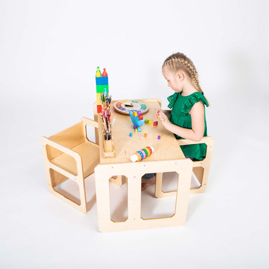 A child in a green dress sits at a Montessori weaning table and 2 chair set, engaging in arts and crafts with colorful blocks, markers, and brushes. This activity fosters fine motor skill development and encourages toddler independence.