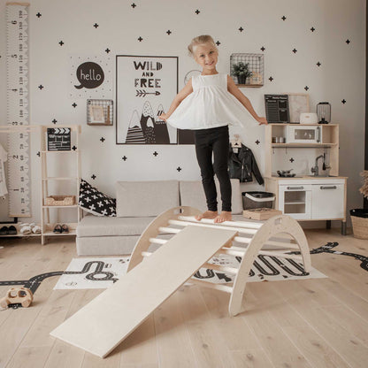 A little girl standing on a Sweet Home From Wood Climbing arch + Transformable climbing cube / table and chair  + a ramp in a room.