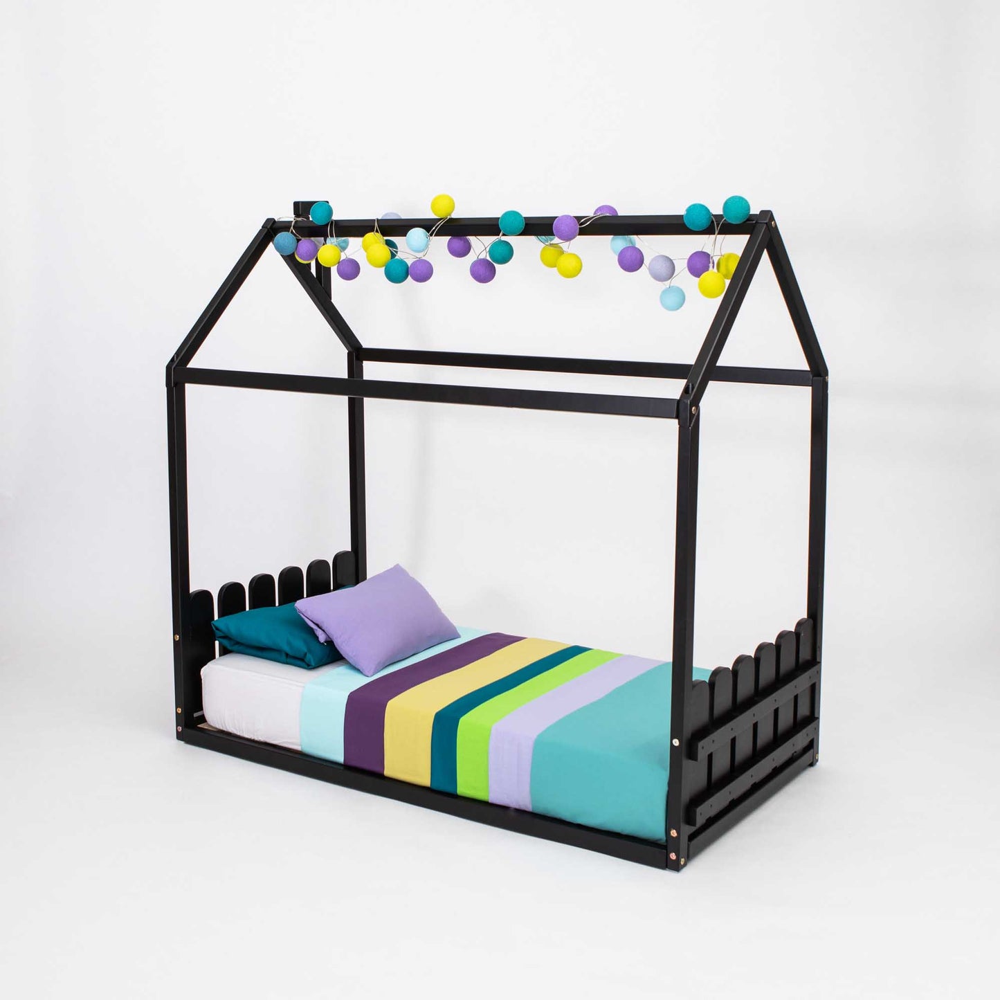 A colorful pom poms-adorned black Montessori house-frame bed with a picket fence headboard and footboard.