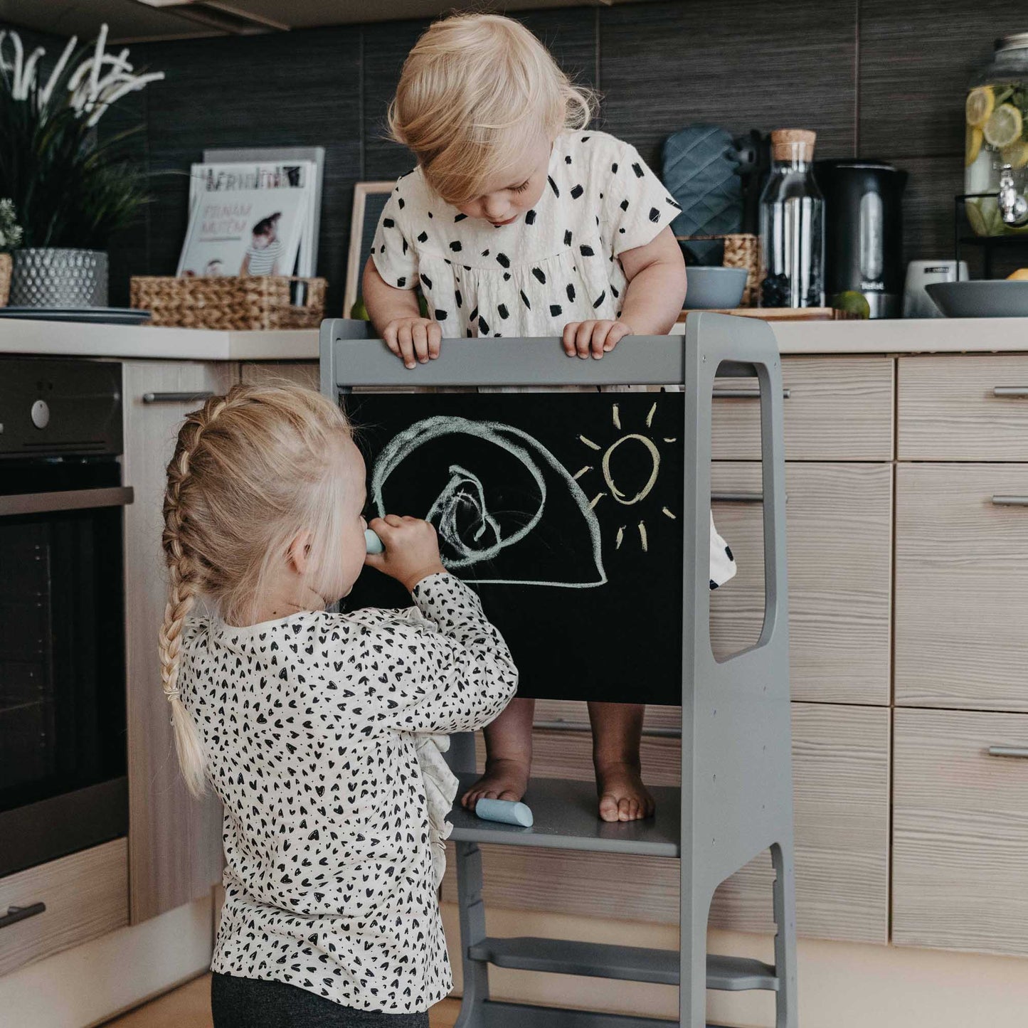 Two little girls using a Sweet Home From Wood Kitchen Tower with Blackboard while playing in a kitchen.