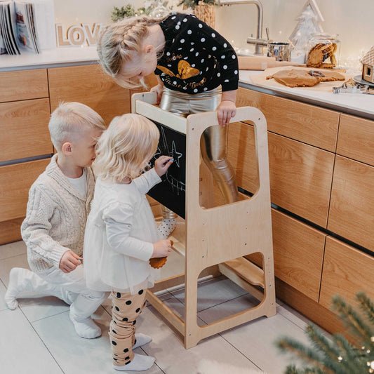 Two children playing with a Sweet Home From Wood kitchen tower with blackboard in a kitchen.