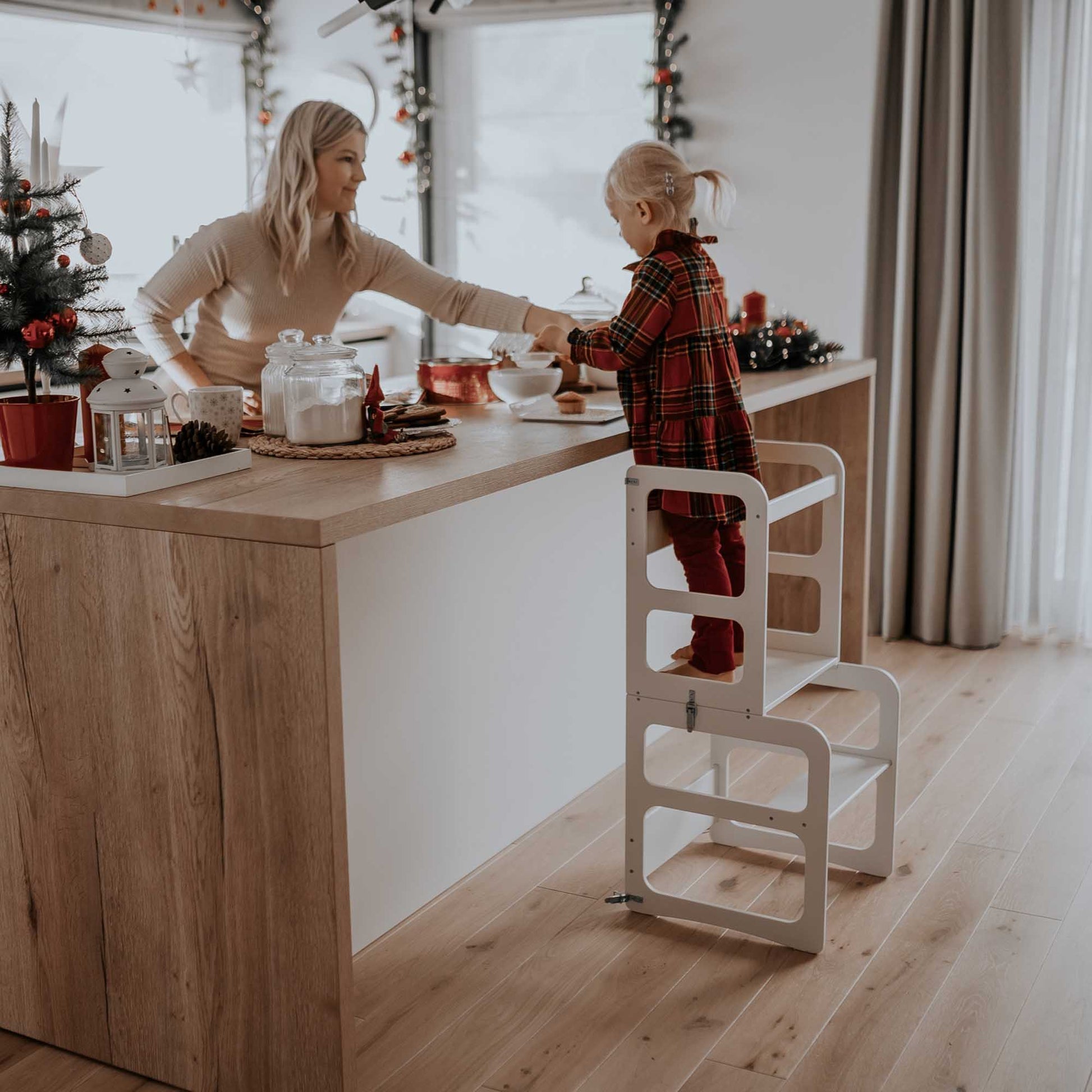 A mother and toddler utilizing a Sweet Home From Wood 2-in-1 transformable kitchen tower - table and chair set in the kitchen.