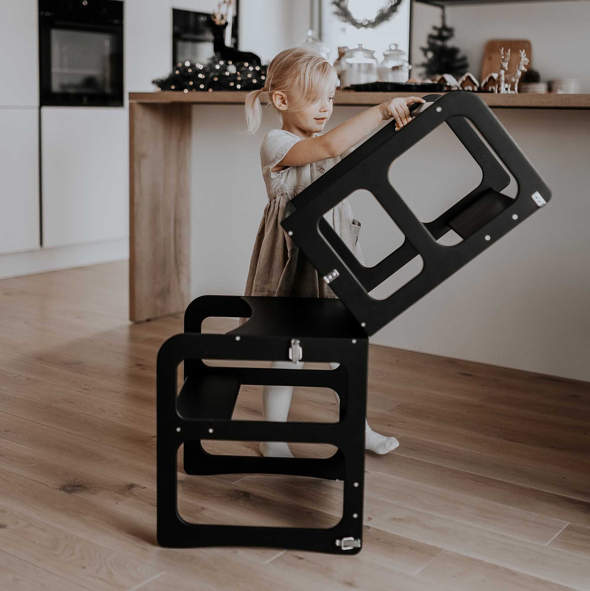 A little girl using a Sweet Home From Wood 2-in-1 transformable kitchen tower - table and chair set in a kitchen.