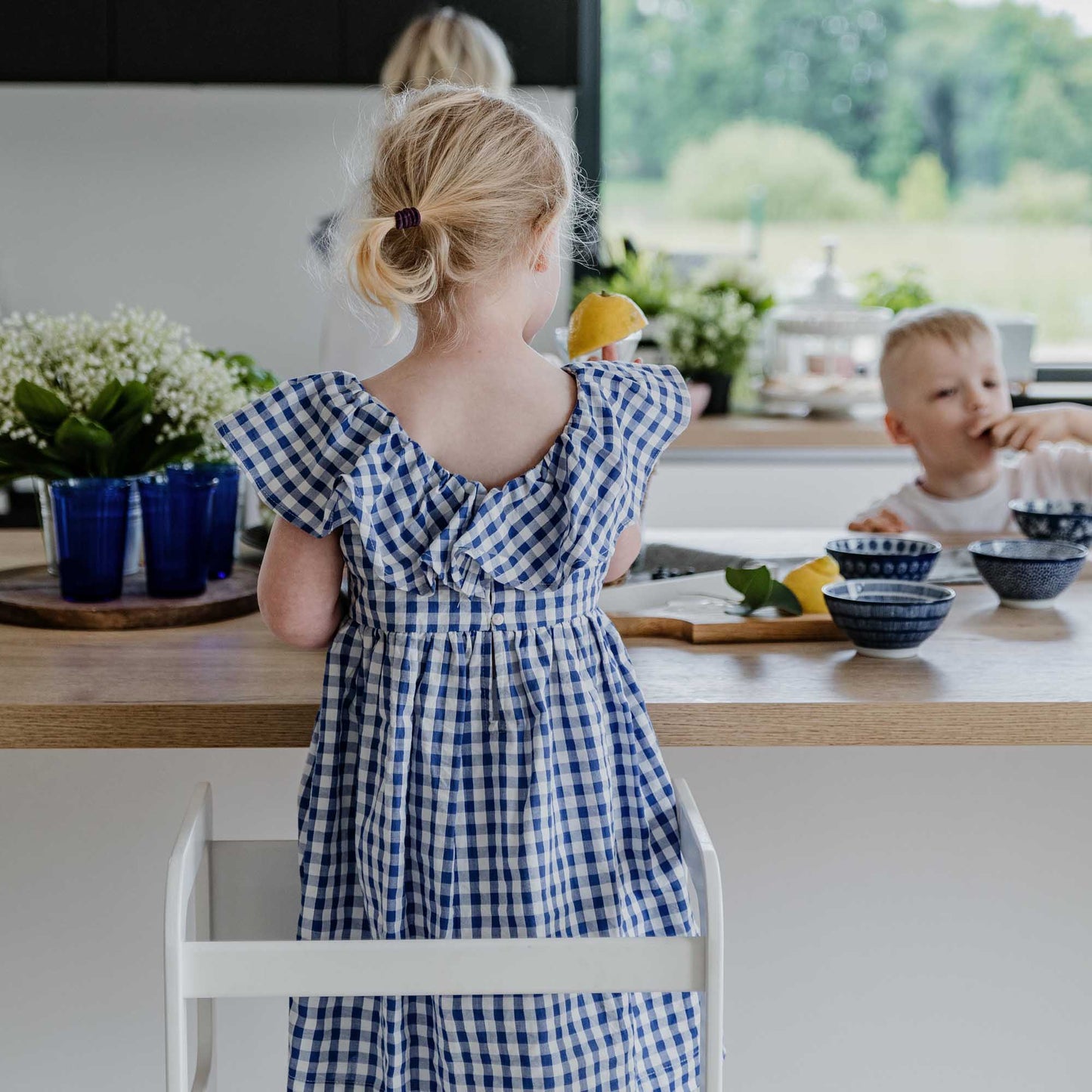 A little girl in a blue gingham dress standing on a Sweet Home From Wood 2-in-1 transformable kitchen tower - table and chair set at the kitchen counter.