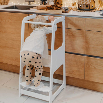 A little girl standing on a Sweet Home From Wood Kids' kitchen tower with 3 height levels in a kitchen.