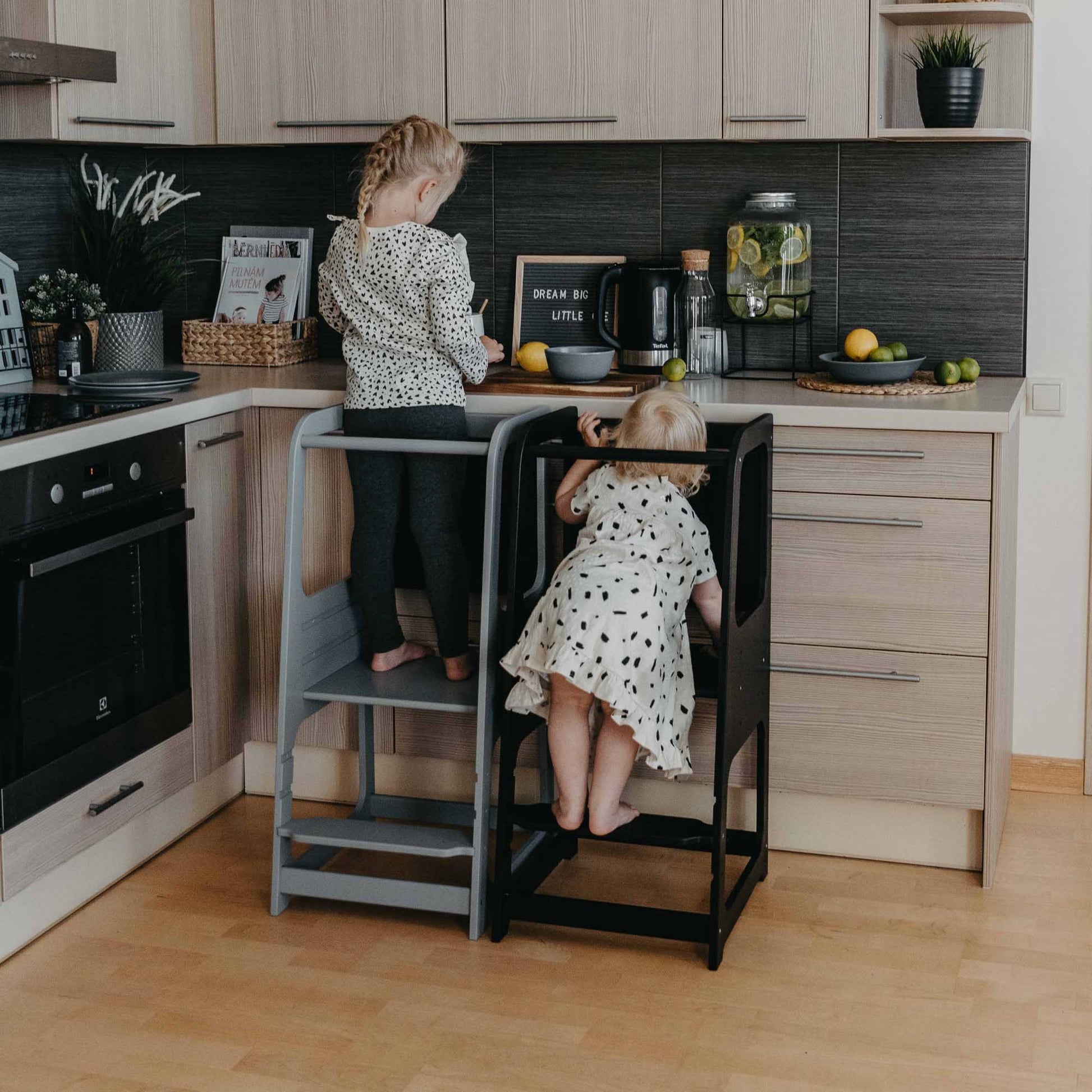 Two siblings standing on a Montessori step stool, joyfully engaged in cooking together. Encouraging independence and family bonding in the kitchen.