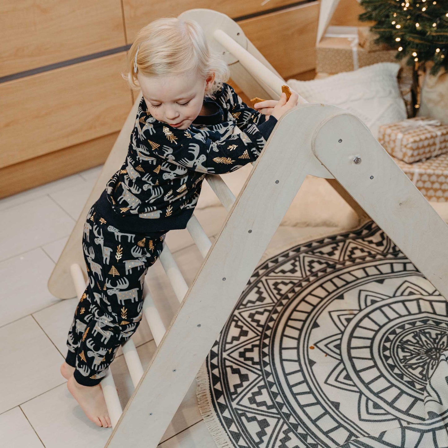 A child is climbing on a Sweet Home From Wood Climbing arch + Transformable climbing triangle + a ramp in front of a Christmas tree.
