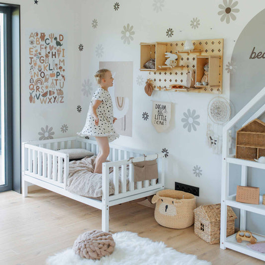A child in a floral dress jumps on a white, floor-level 2-in-1 toddler bed on legs with a vertical rail fence in a bedroom decorated with flower decals, an alphabet chart, and wooden shelves holding decor items. Baskets and toys sit on the floor by the solid wood bed.