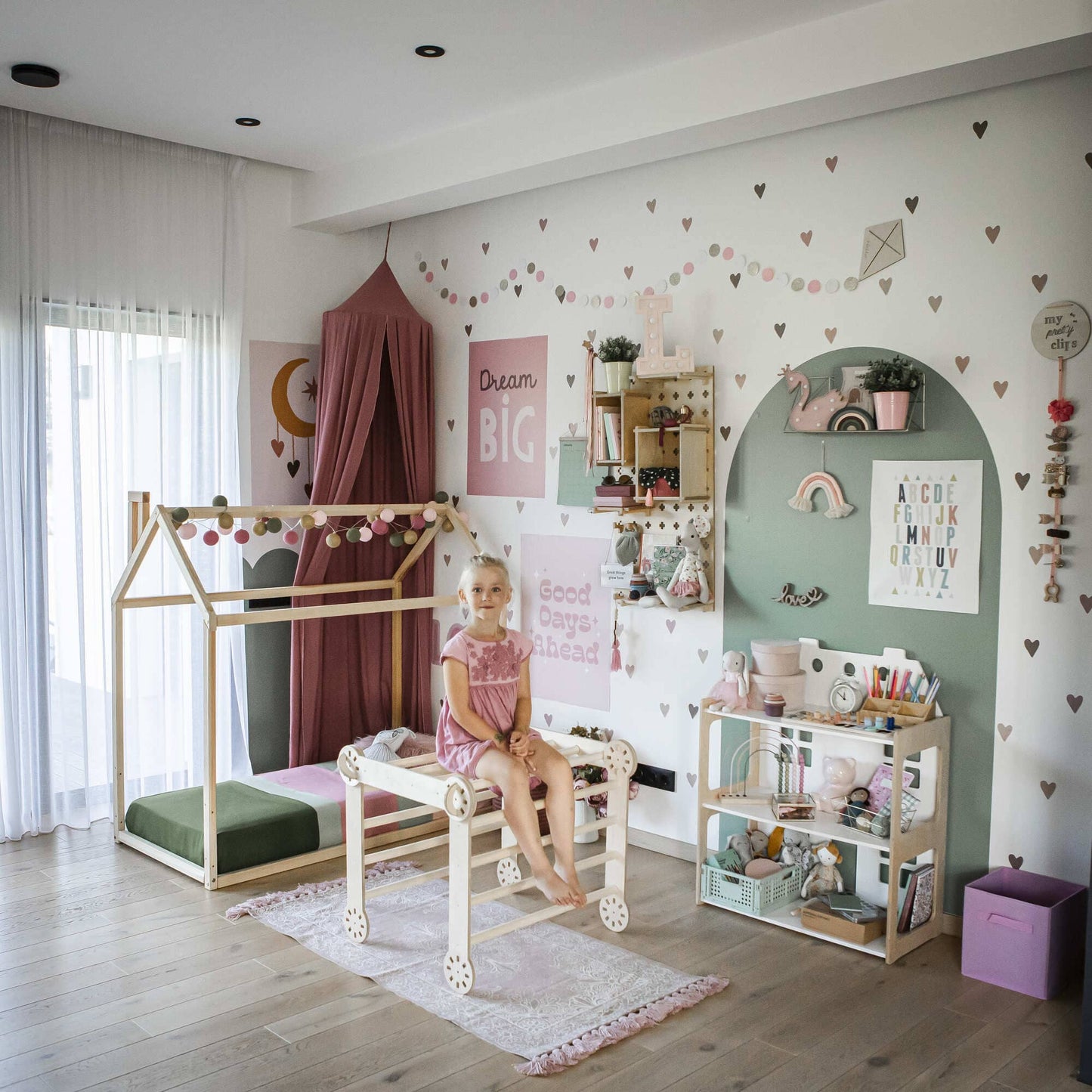 A young girl in a pink dress sits on a wooden horse-shaped indoor toy in a children's room, which features an activity gym called the Transformable Climbing Gym, a house-shaped bed, shelves, and wall decorations adorned with motivational quotes and heart stickers.