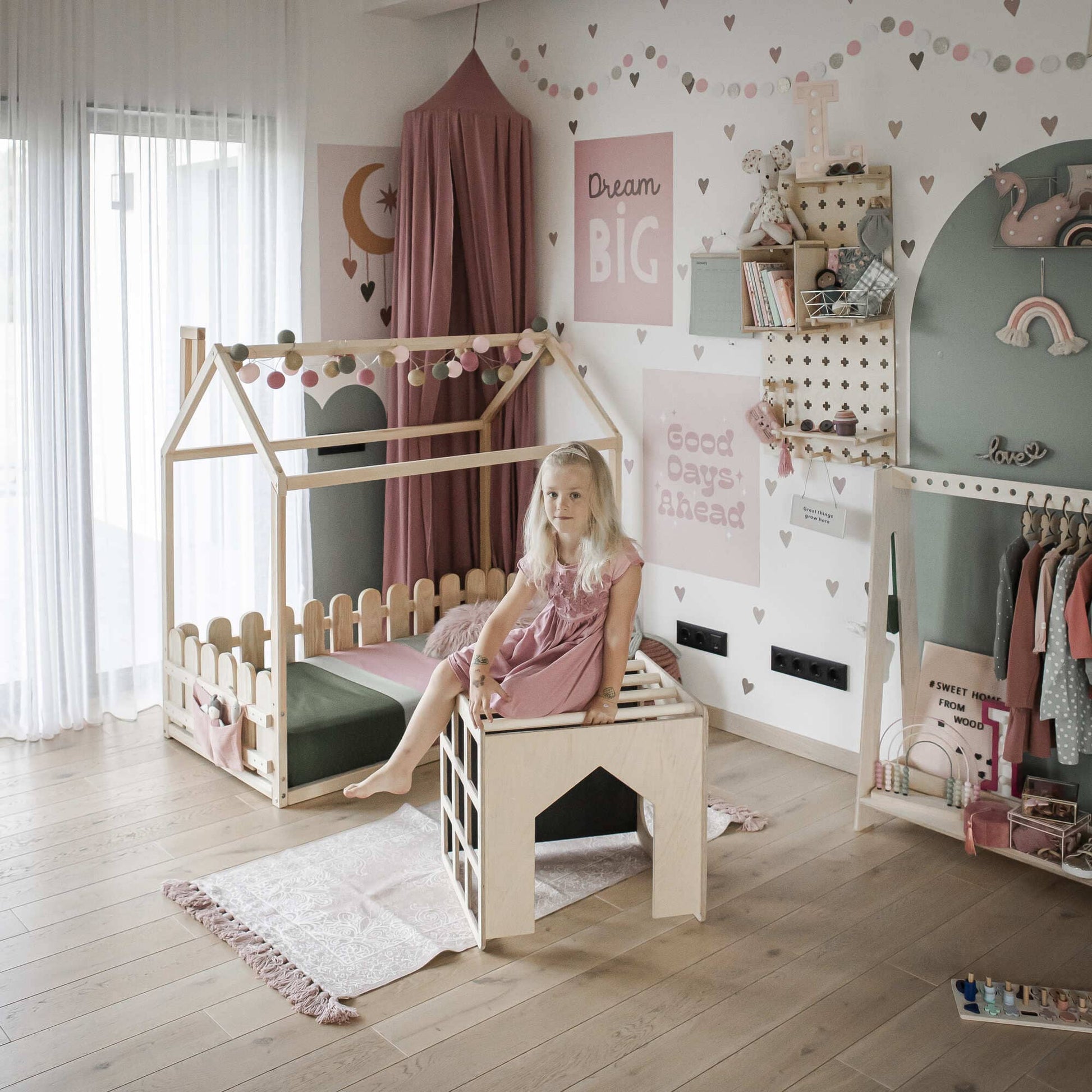 A young girl sits on a small chair in a neatly arranged children's room with a wooden playhouse, decorative wall art, and various toys organized around the space, including a Montessori table and an activity cube with sensory panels.