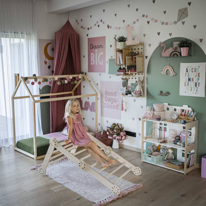 A young girl sits on a Transformable climbing gym in a well-decorated children's room featuring an activity gym, a bed shaped like a house, various wall decorations, and shelves filled with indoor toys and books.