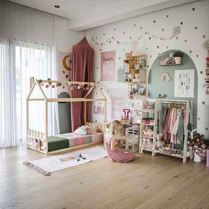 Child's bedroom with a canopy bed, wall art, a reading nook, toy shelves, and a Montessori children's wardrobe crafted from Baltic birch plywood. A child sits on the floor playing near a rug with toys. The room is decorated with hearts and pastel colors featuring the Children's Wardrobe and Dress-Up Clothing Rack.