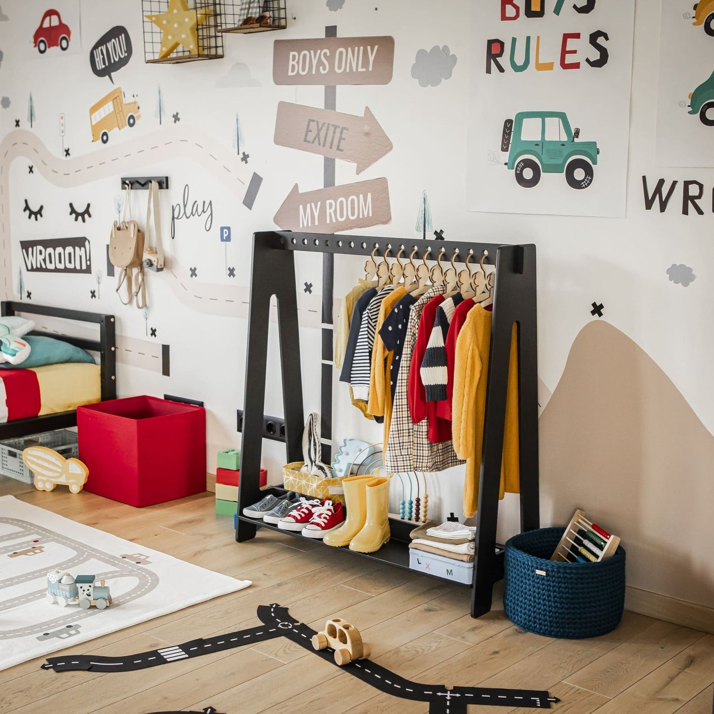 A children's bedroom featuring a Montessori clothing rack adorned with colorful clothes, toys neatly arranged on shelves, and a play rug displaying road patterns. The walls are decorated with whimsical decals, and the room boasts wooden flooring designed for optimal closet organization.