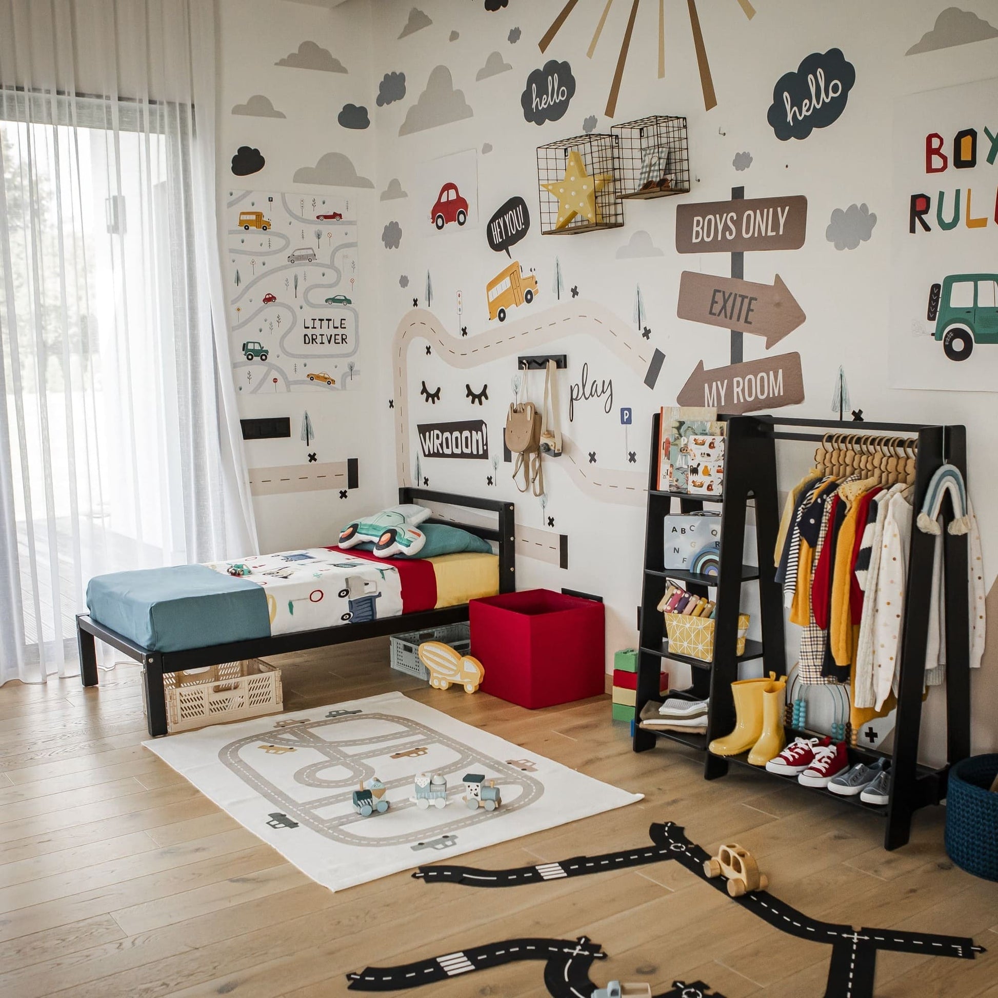 A children's bedroom with car-themed decorations features a single bed, a children's wardrobe and dress-up clothing rack crafted from Baltic birch plywood, colorful outfits hanging neatly, storage bins, and a playmat with toy vehicles scattered on the floor.