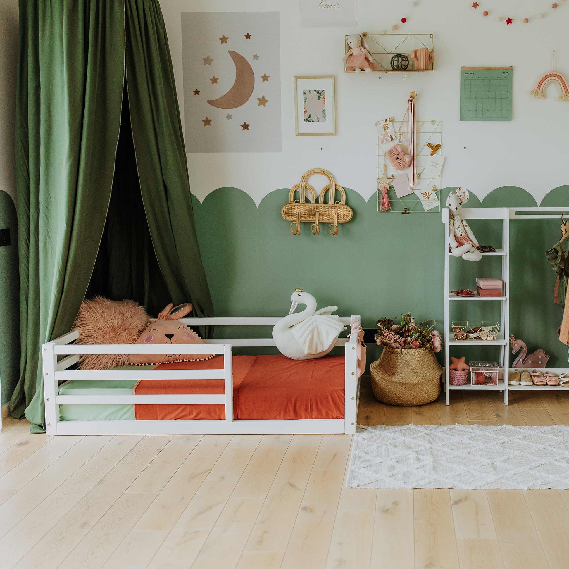 A children's bedroom with a white, floor-level kids' bed featuring a horizontal rail fence and a solid wood frame, surrounded by green curtains. A swan plush toy and various wall decorations add charm, while a shelf and closet organizer hold toys and clothes, promoting independent sleeping.