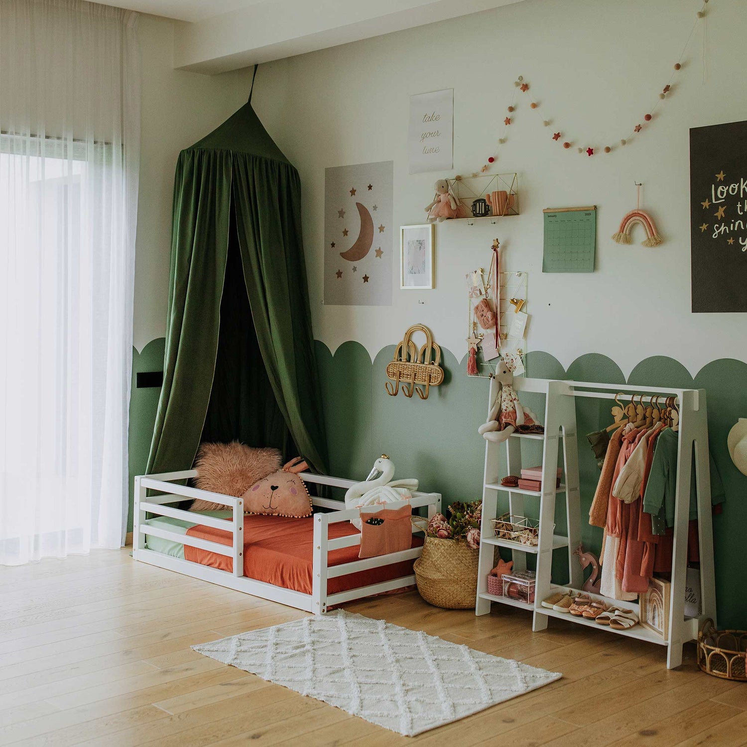 A child's bedroom featuring a floor-level kids' bed with a horizontal rail fence, dressed in orange bedding, accompanied by a green canopy. The room is adorned with various wall decorations, shelves filled with toys, and a clothing rack. The pale green and white color scheme creates a serene space filled with natural light to encourage independent sleeping.