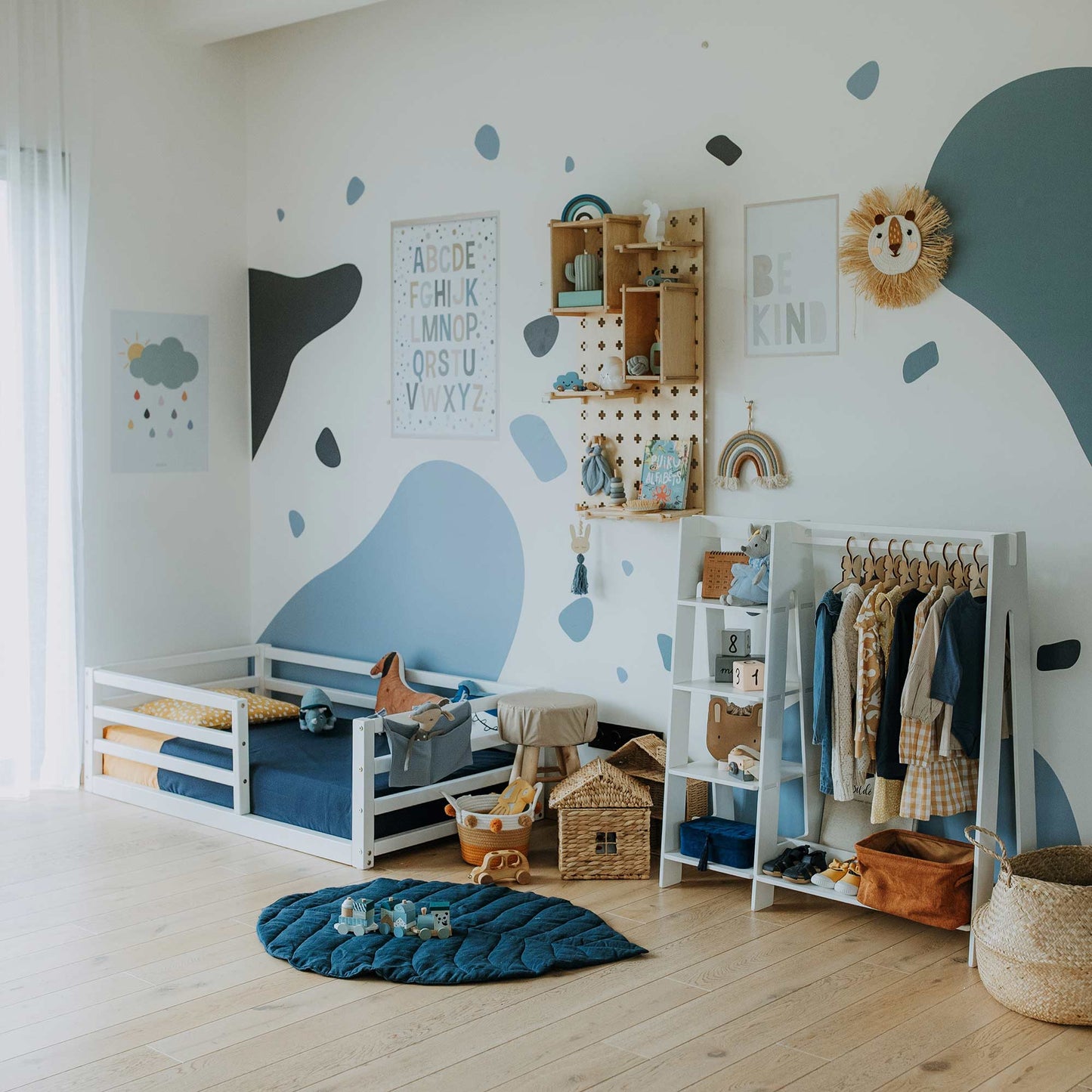 A children's room outfitted with a floor-level kids' bed featuring a horizontal rail fence designed for independent sleeping, along with a toy shelf, a clothing rack, and wall decorations including letters, a rainbow, and a lion. The room boasts a blue and white color scheme accented with polka dots.