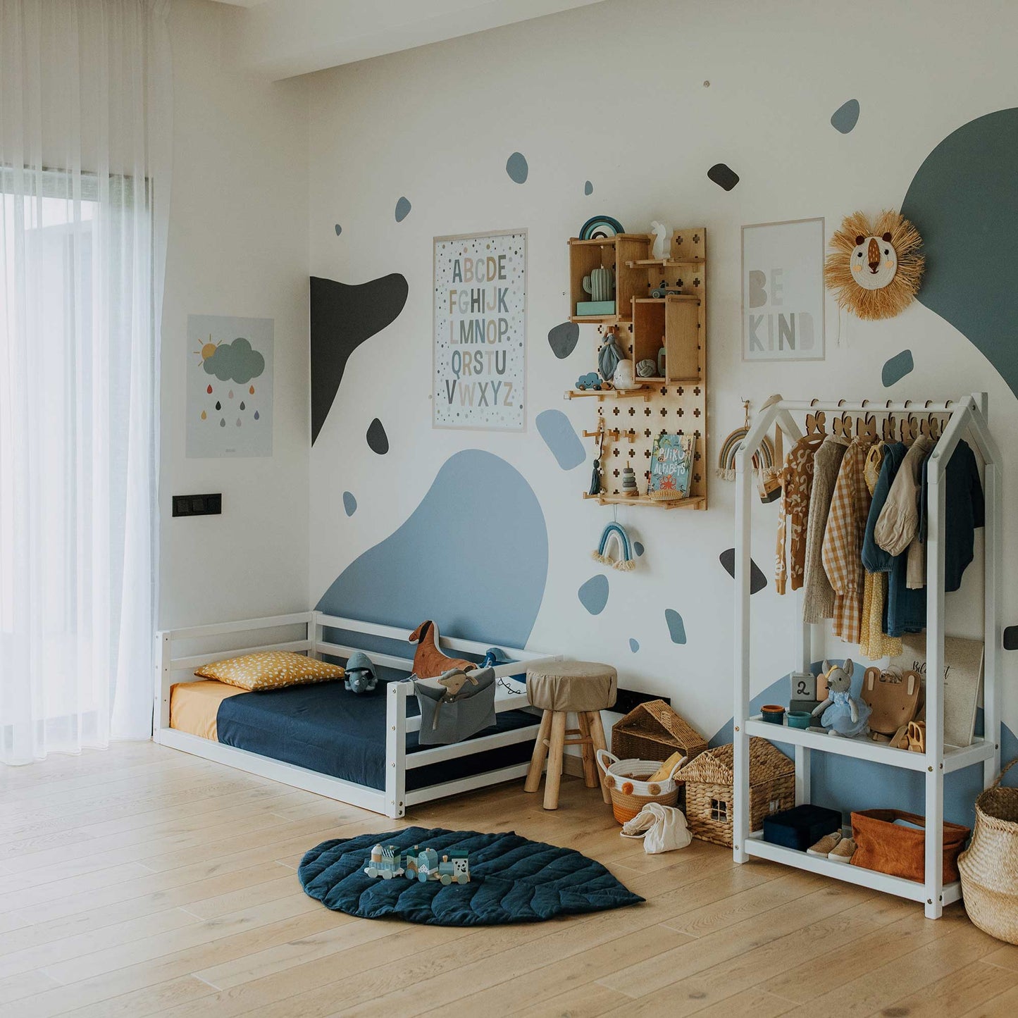 A children's bedroom with a blue and white theme features a Children's floor level bed with a 3-sided safety rail and solid pine wood frame, shelf with books and toys, clothing rack, and wall decor. Various storage baskets and a play mat are also visible.